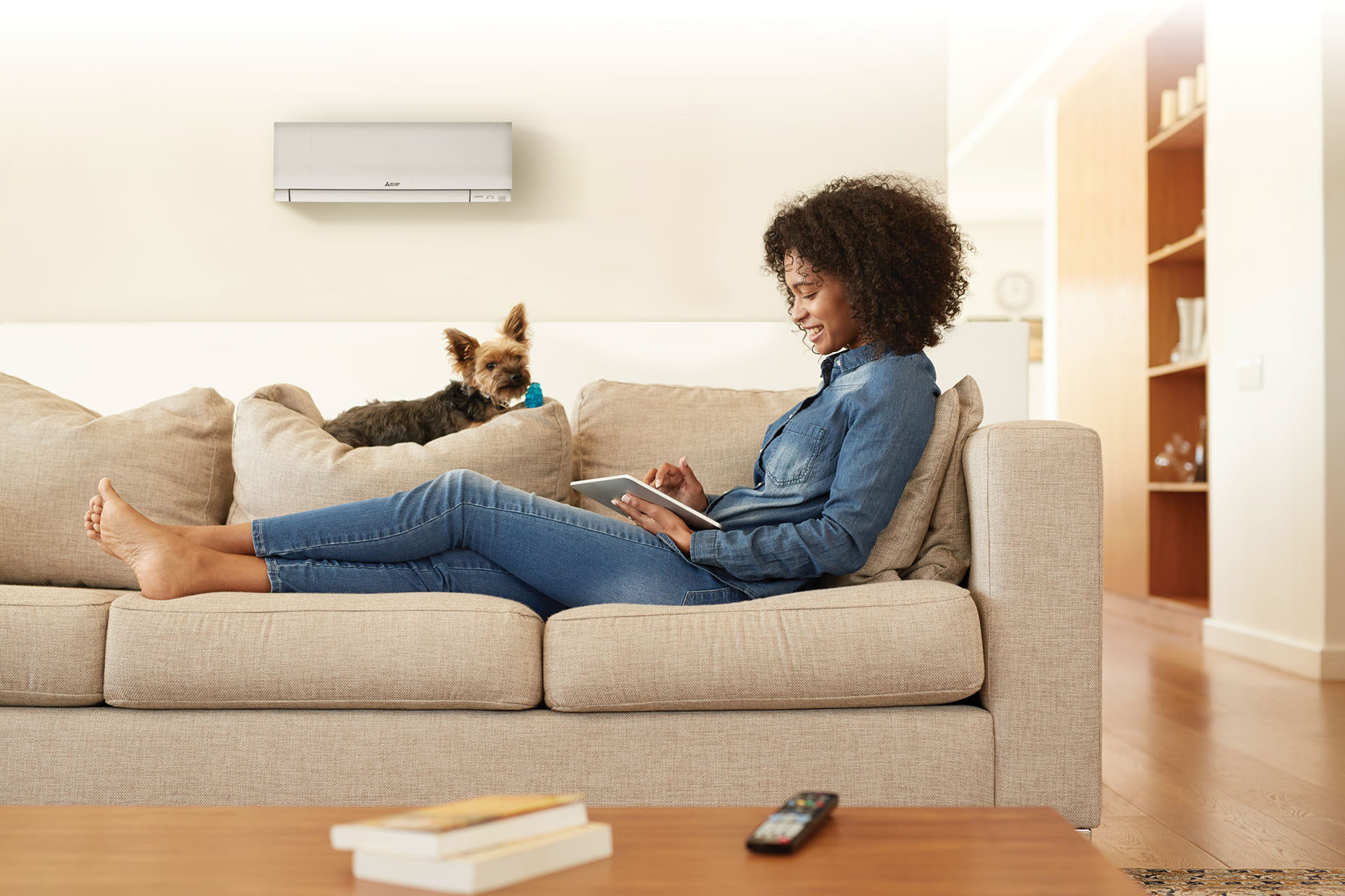 A woman sits on a sofa with her dog with a Mitsubishi ductless air conditioning and heating system on the wall behind them.
